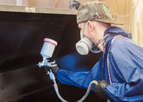 Operario con mascarilla y traje azul aplicando lacado con pistola de spray sobre mueble oscuro en taller en Burgos