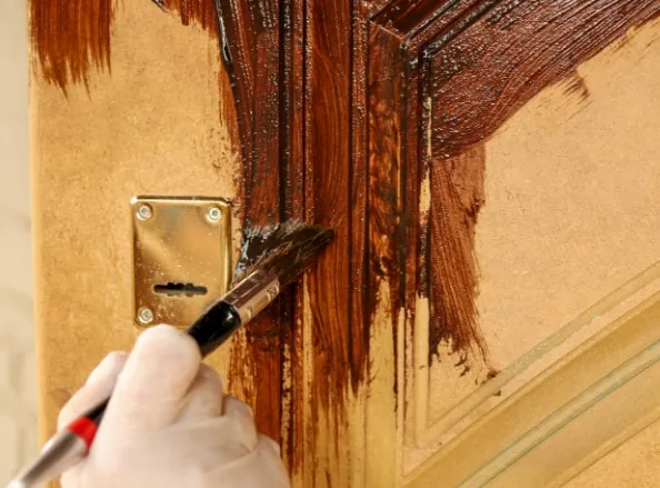 Mano aplicando barniz oscuro con brocha sobre puerta de madera tallada junto a bocallave dorada en Burgos