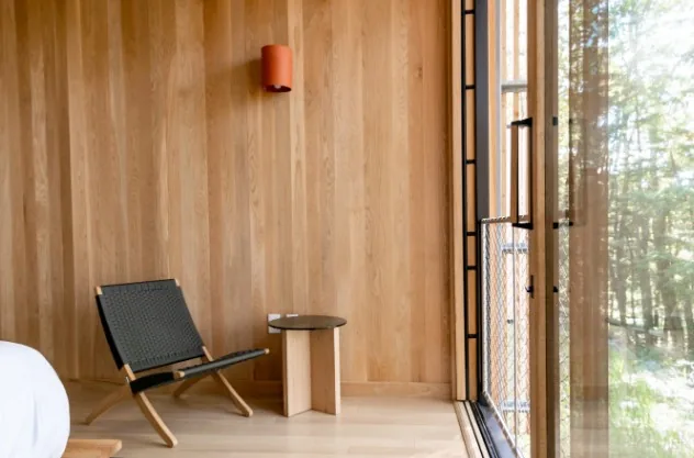 Interior de cabaña con paredes revestidas de madera clara, silla negra, mesita y puerta acristalada al bosque en Burgos